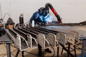 Welder in protective helmet and jacket welding metal frames on a workbench, with an exhaust hose and scattered tools nearby.