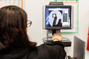 Technician operating an eye-diagnostic machine; hand on the control panel with a monitor showing a circular target and data readouts.