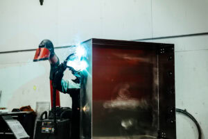 Welder in protective gear welding a large metal cabinet with bright sparks flying from the weld point.