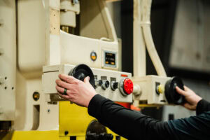 Worker operating a beige industrial machine, turning large black control discs on a factory panel.