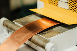 Copper sheet on a rolling machine, shiny orange-brown metal passing under a yellow guard plate.
