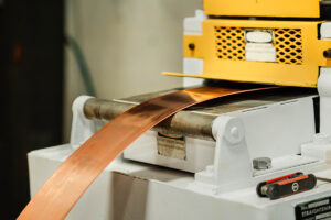 Copper strip being fed through a yellow metalworking roller machine on a white base, shaping the copper sheet in a factory setting.