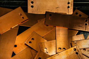 Pile of copper-colored metal plates with circular holes, stacked chaotically in warm lighting (industrial scrap).