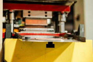 Yellow industrial machine with a metal press area and tooling visible on top.','Close-up of a stamping press inside a yellow safety guard showing dies and rails.','Industrial manufacturing equipment: yellow enclosure surrounding a metal die area with precision tooling.','View of a yellow press cabinet with metallic components and tooling extending above the guard.','Sturdy yellow safety housing around a metal stamping machine’s tooling region in a factory setting.