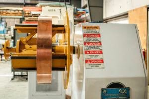 Industrial metal forming press with a large copper band and yellow frame in a workshop, safety labels on the side panel рядом.