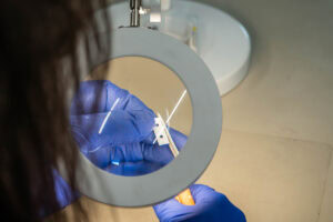 Person wearing blue gloves inspects a small white bracket with holes under a circular magnifier in a lab setting