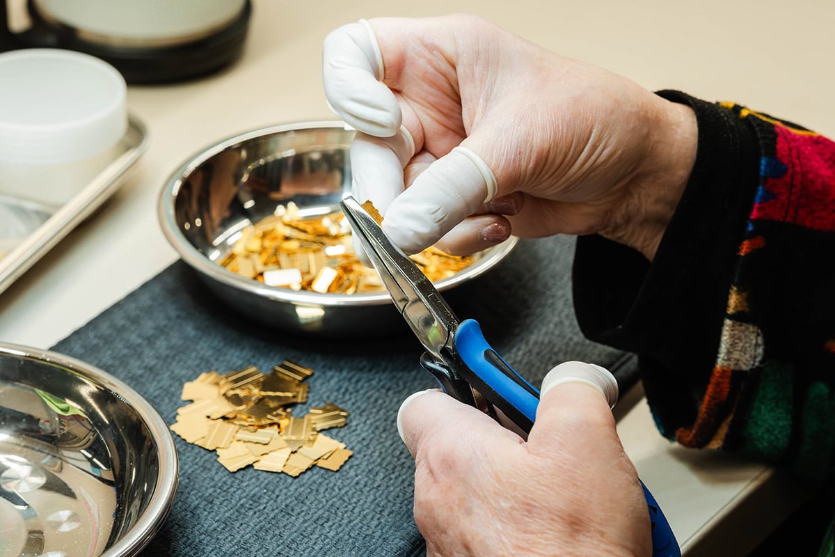 Gloved hands trim gold foil sheets with pliers over a metal tray of gold pieces for crafting/jewelry making