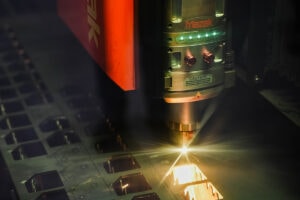 Industrial laser cutter in action, sparks flying as a laser head cuts a metal sheet, Mazak logo visible on the machine.