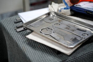 Stainless steel medical instrument tray with cutouts, resting on a textured dark table with towels and folders nearby.