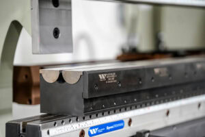 Industrial metal stamping press with a black rectangular die holder mounted on a precision bed and a ruler scale visible at the front.