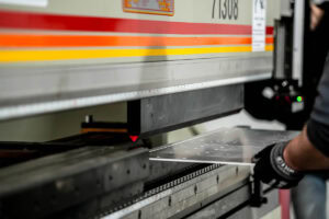 Worker wearing gloves guiding a sheet of metal under a large industrial machine.