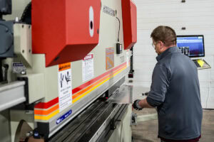 Worker wearing safety glasses operates a large metal press brake, guiding a sheet metal piece in a workshop.