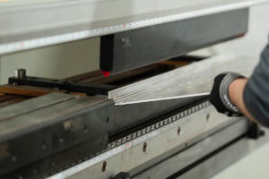 Technician wearing gloves feeds a sheet of metal into a press brake for bending.