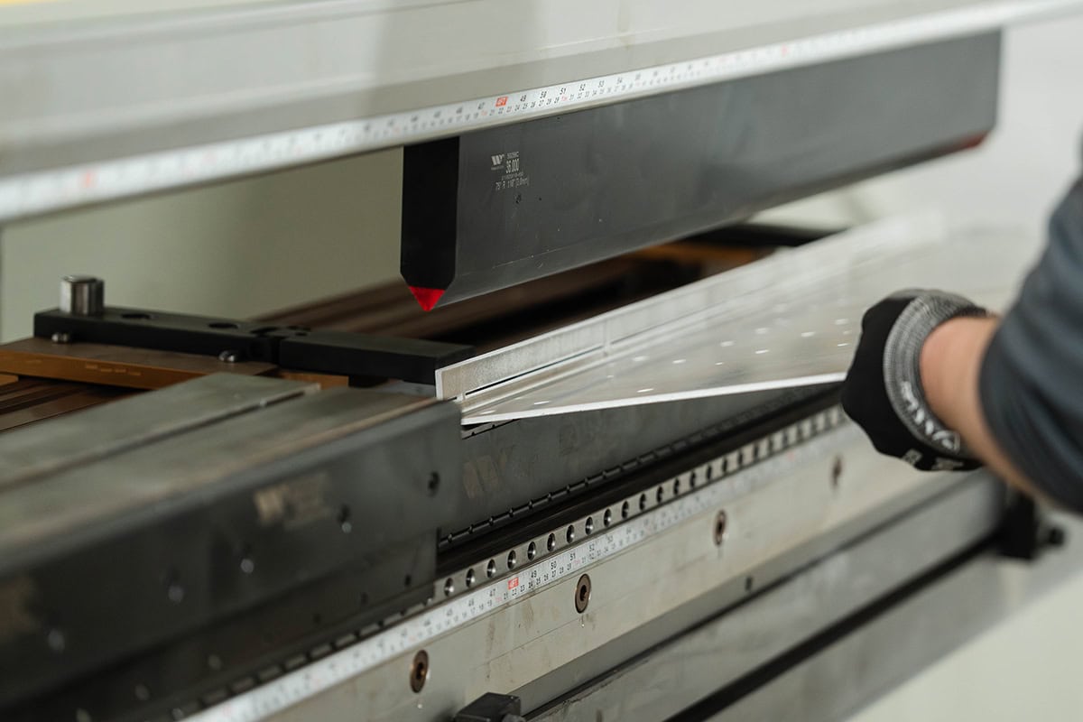 Technician wearing gloves feeds a sheet of metal into a press brake for bending.