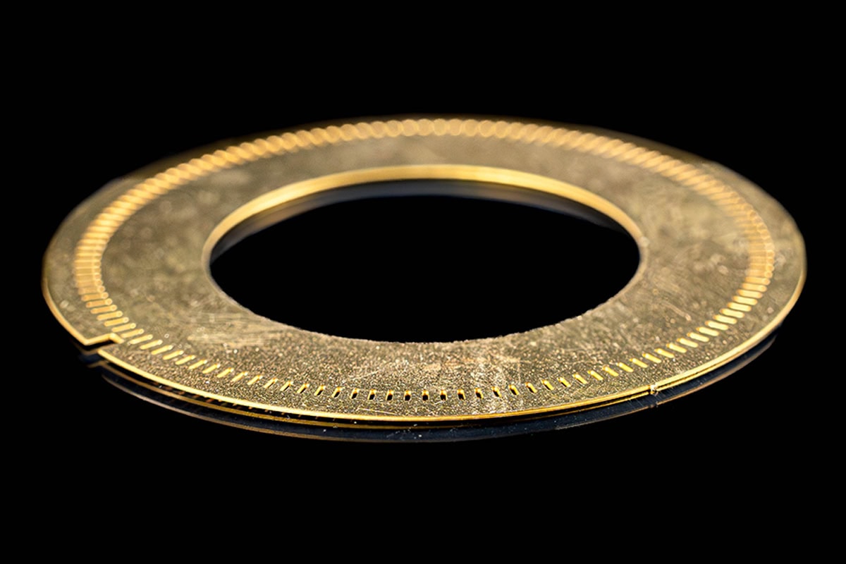 Close-up of a gold circular ring with evenly spaced tiny holes along the inner edge, set against a black background.