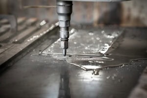 Close-up of a drill bit cutting into a metal plate in a workshop, with coolant pooling around the work area.