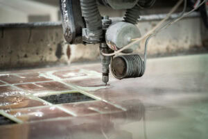 Industrial waterjet cutting head engaged on metal tiles in a workshop, with water pooling on the surface.