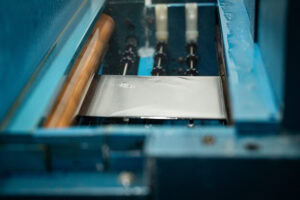 Metal sheet moving through a blue industrial machine on a conveyor or press system.