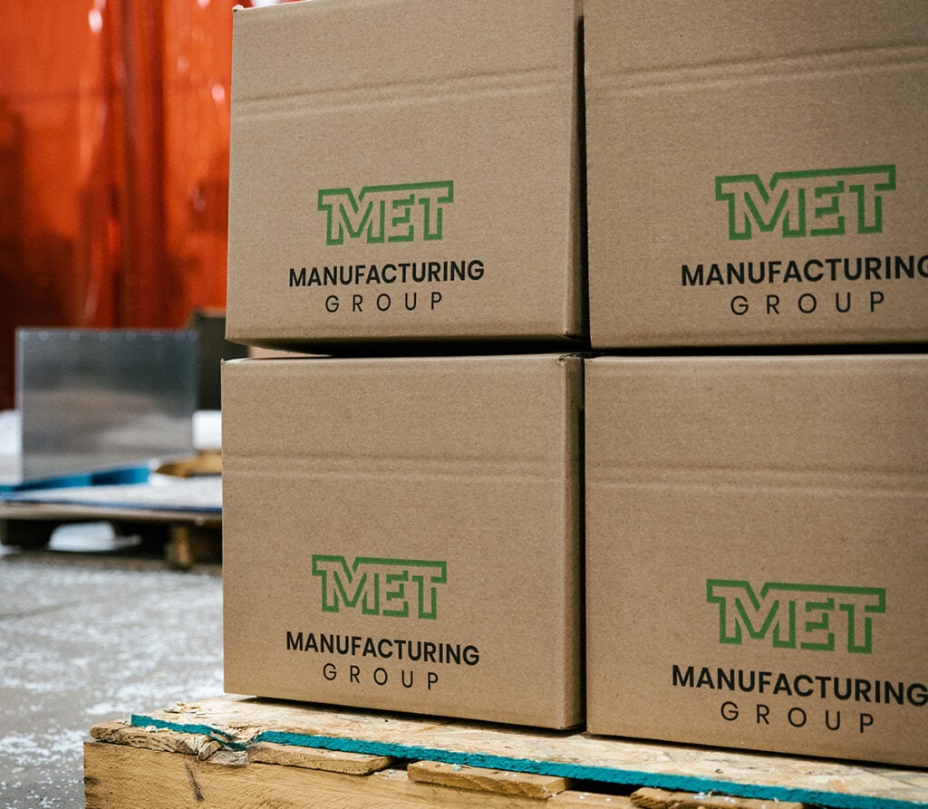 Stack of brown cardboard boxes with a green MET Manufacturing Group logo on a wooden pallet in a warehouse setting