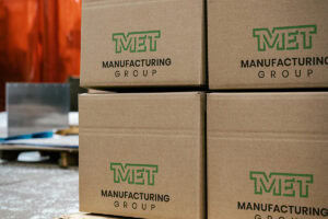 Stack of brown cardboard boxes with a green MET Manufacturing Group logo on a wooden pallet in a warehouse setting