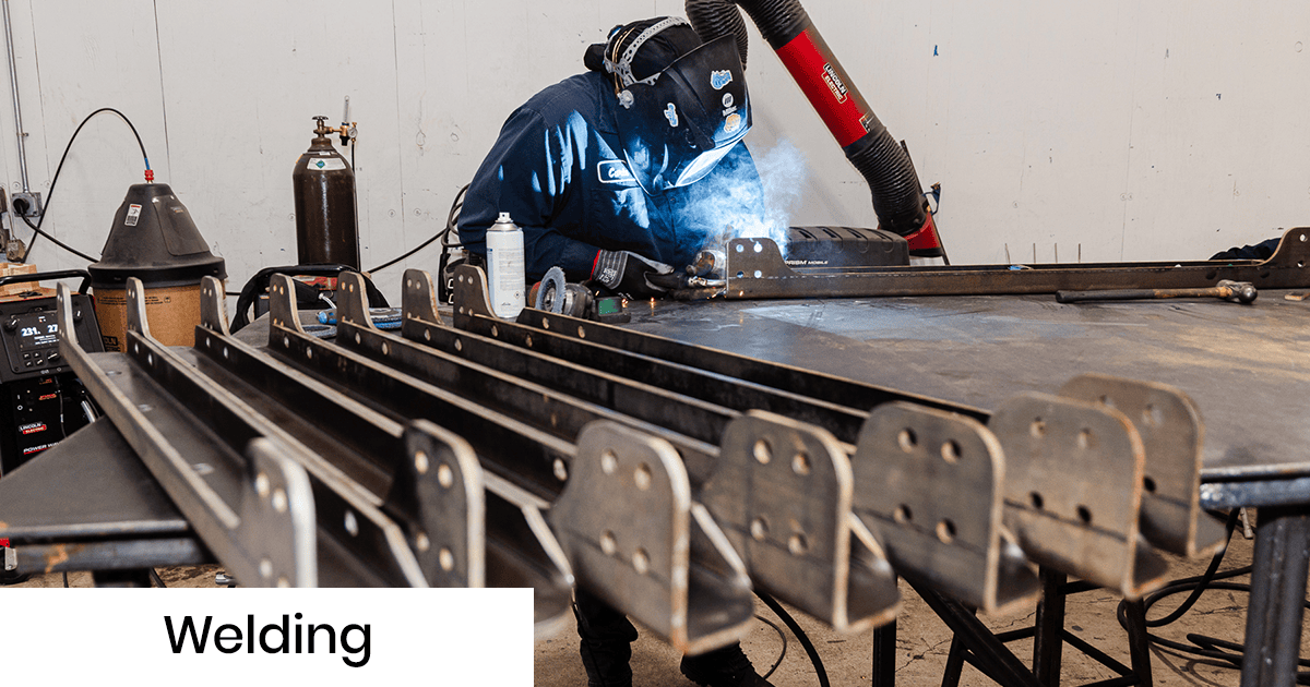Welder in protective gear welding metal beams on a worktable in a shop, with welding hose and equipment nearby.